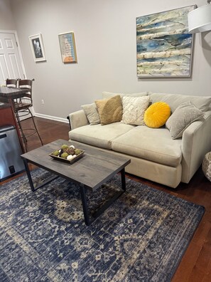 A living room with a beige sofa, a coffee table with decorative items, a patterned rug, and a painting on the wall. -     Charming 2BR Baltimore Retreat Near Johns Hopkins (Baltimore)