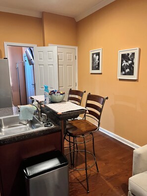 A kitchen with a sink, chairs, and a trash can. There are two doors and a wall with framed pictures. -     Charming 2BR Baltimore Retreat Near Johns Hopkins (Baltimore)
