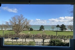Blick aus einem Fenster auf einen Park mit Bäumen, ein Gewässer und eine entfernte Küste.