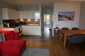 A compact living space with a kitchenette, a dining area with a table and chairs, a red sofa, and a bookshelf. - Holiday apartment with 1 bedroom (Lübeck-Travemünde)