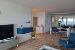 A modern living room with a flat-screen TV, a blue accent wall, a dining area with a table and chairs, and a view of the sea through a glass door. - Holiday apartment MeerZeit (Timmendorfer Strand)