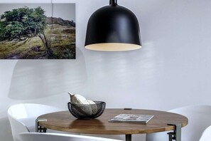 Une table ronde en bois, surmontée d’un bol en métal noir et agrémentée d’un journal, une lampe suspendue noire et des chaises blanches, dans une pièce moderne au design minimaliste.
