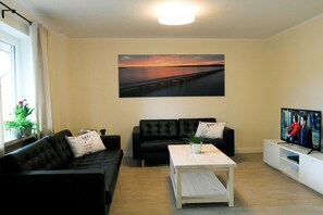 A living room with a black sofa, a white coffee table, and a TV on a white stand. - Holiday apartment with 1 bedroom (Fehmarn OT Burg)