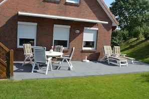 A brick house with a patio area featuring a table and chairs, a covered seating area, and a view of a grassy hill. - Holiday home with 3 bedrooms (Neßmersiel)