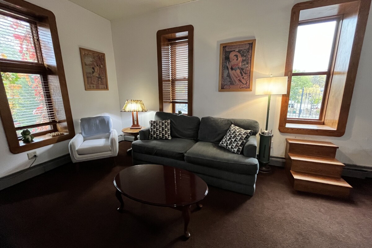 A living room with a sofa, armchair, and coffee table, featuring large windows and a staircase.