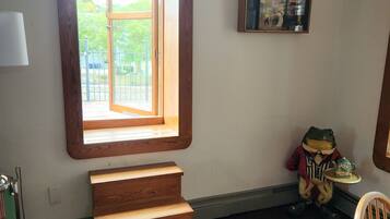 A room with a wooden staircase, a window with a view of greenery, a framed picture on the wall, and a small table with a frog figurine.