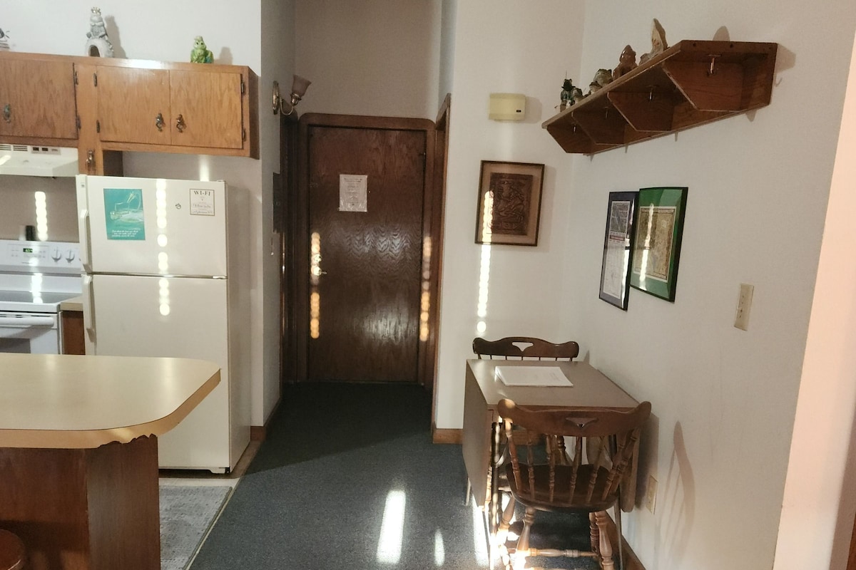 A kitchen with a refrigerator, stove, and wooden cabinets. There is a dining table with chairs and a wall-mounted shelf above it.