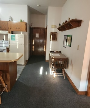 A kitchen with a refrigerator, stove, and wooden cabinets. There is a dining table with chairs and a wall-mounted shelf above it.