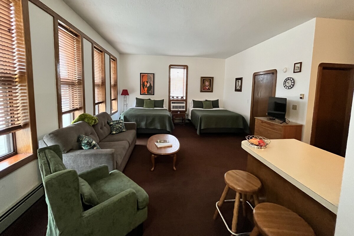 A compact hotel room with a bed, two single beds, a sofa, a small table, and a TV.