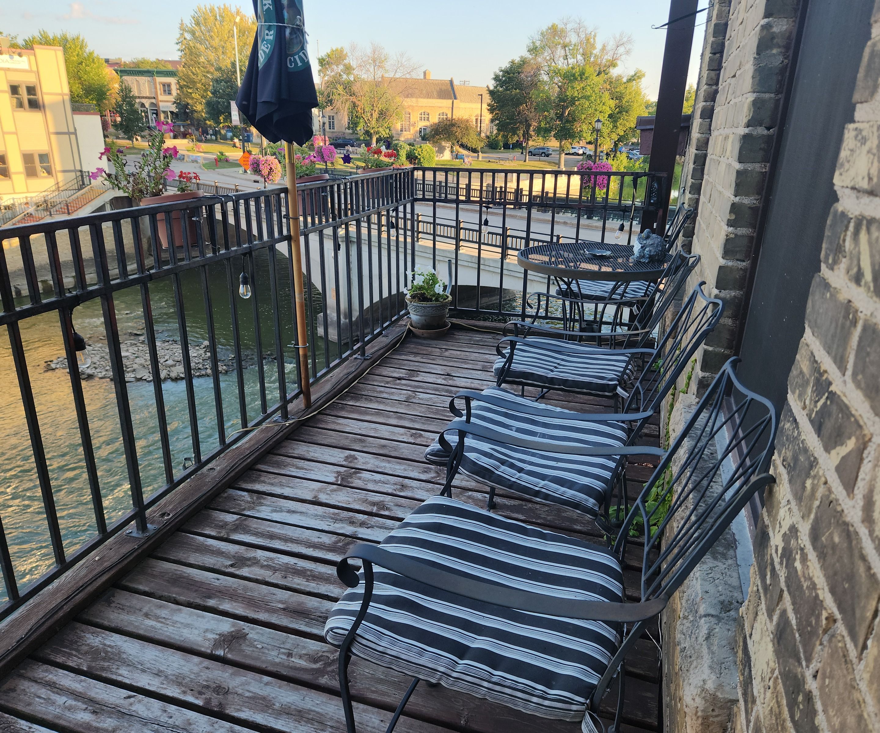 Una terraza de madera con barandillas de metal, una mesa con sillas y una vista de un río y edificios al fondo.