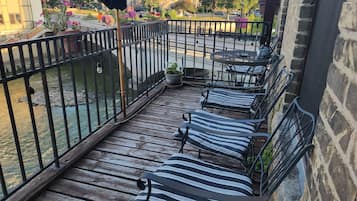 Una terraza de madera con barandillas de metal, una mesa con sillas y una vista de un río y edificios al fondo.