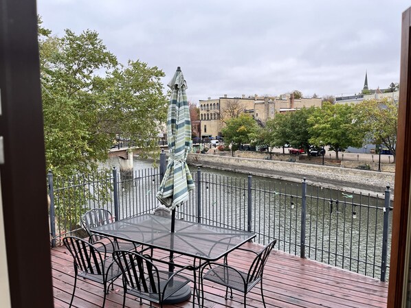 A deck with a table and chairs overlooking a river, with a view of buildings and a bridge in the background.
