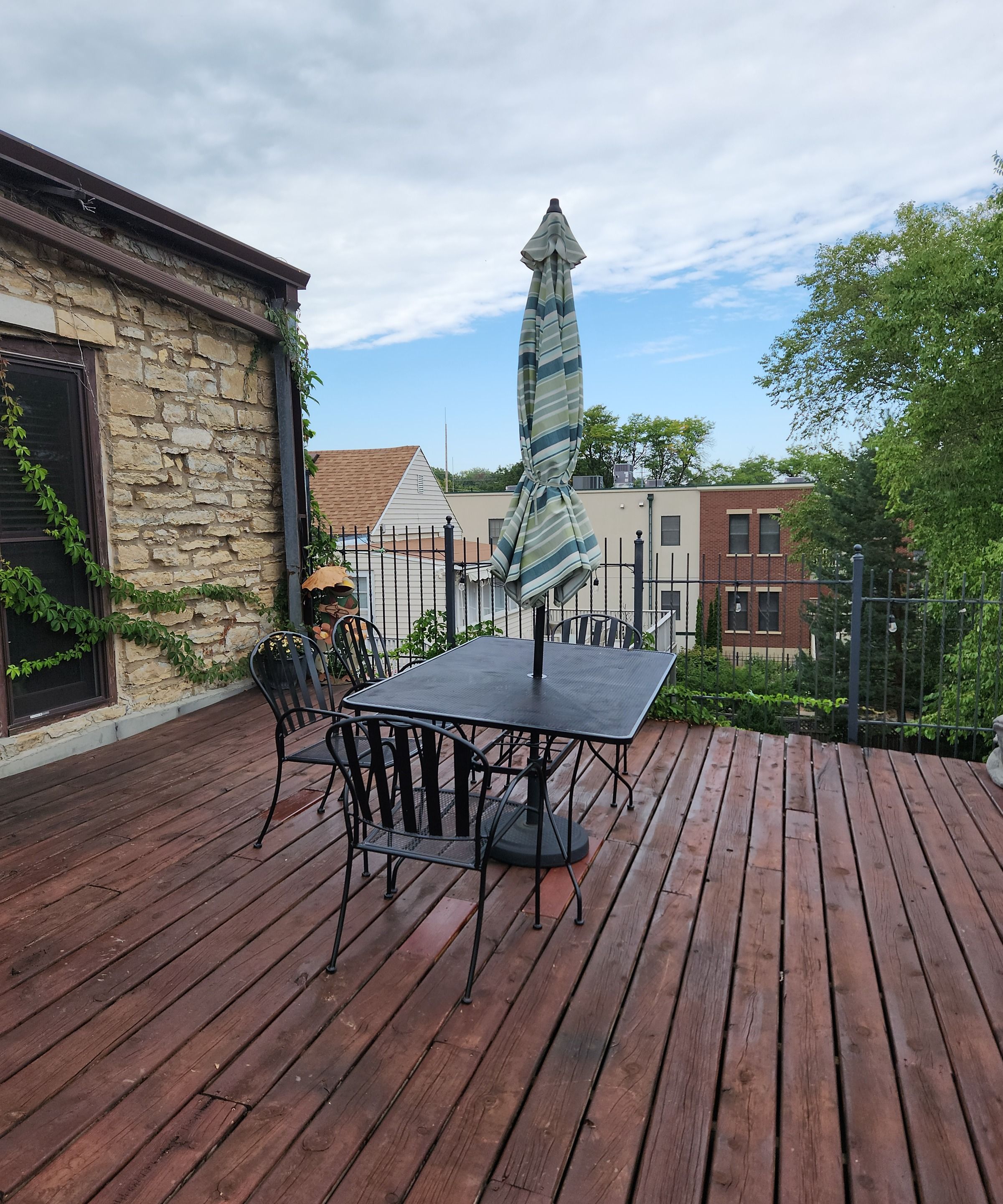 Una terraza de madera con una mesa y sillas, un edificio de piedra y una valla metálica.