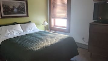 A bedroom with a bed, a nightstand, a lamp, a window with blinds, a television, and a framed picture on the wall.