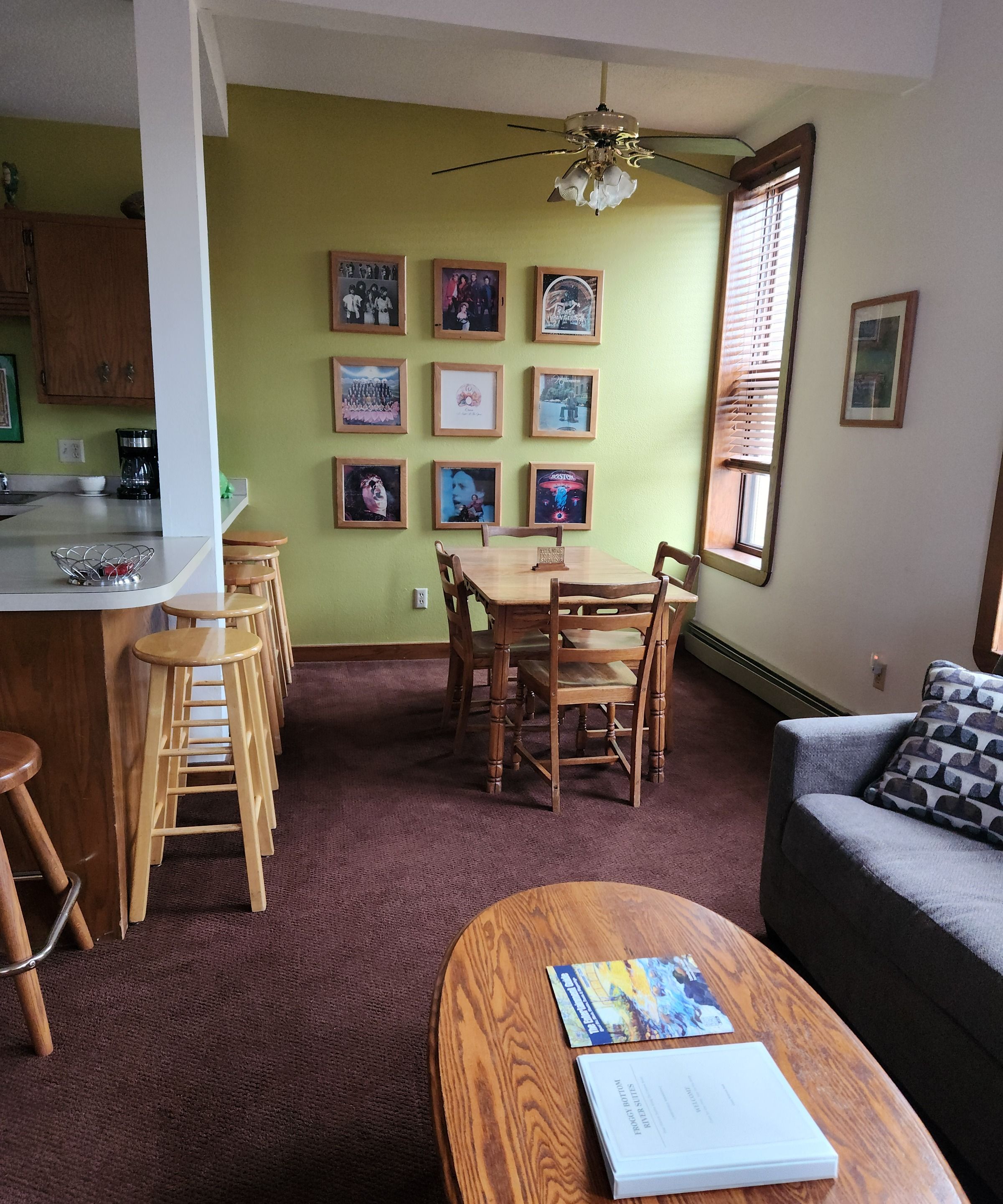 A living room with a dining area, a kitchen, and a ceiling fan. There are framed pictures on the wall and a book on the coffee table.