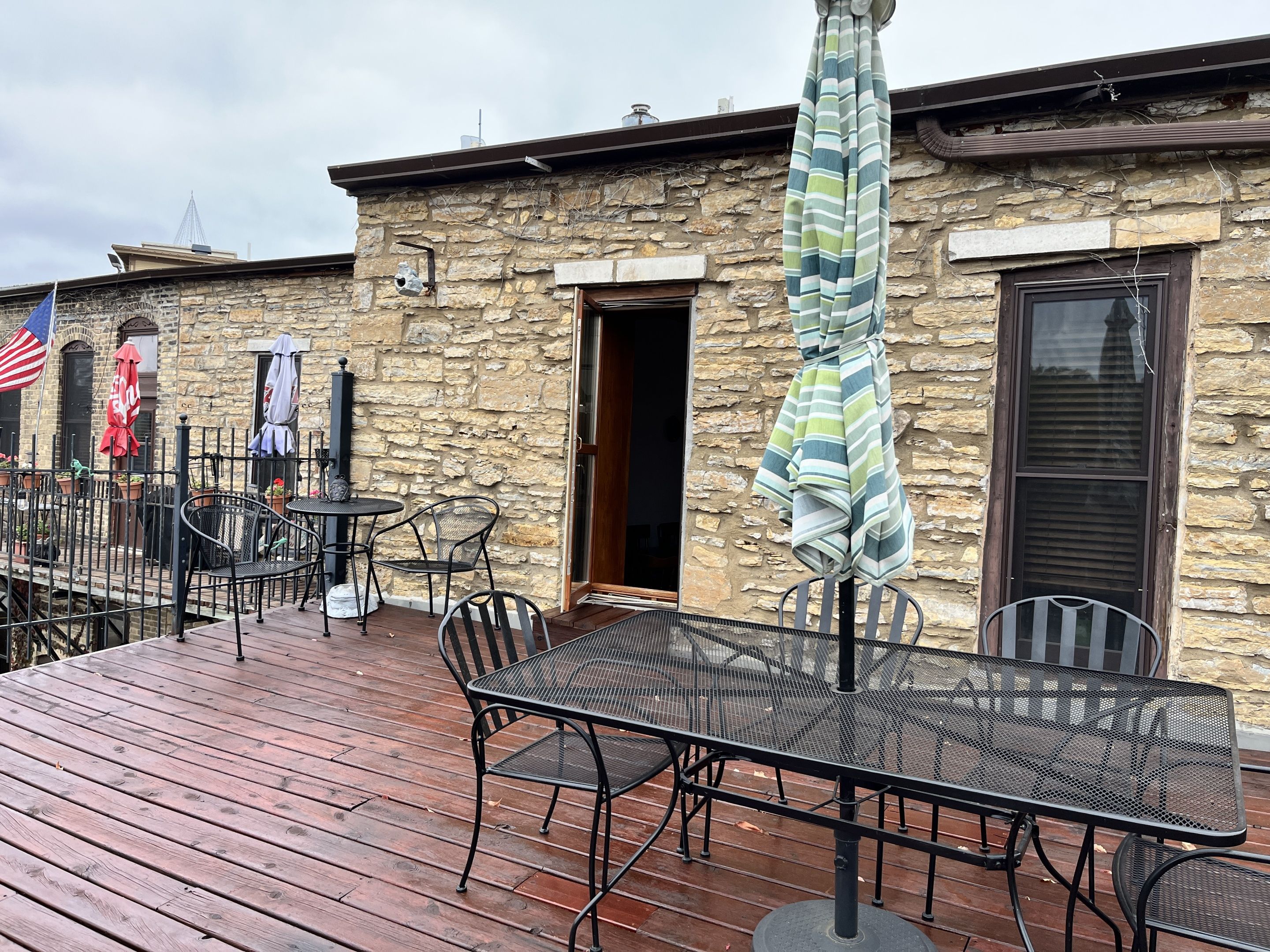 An outdoor patio with metal tables and chairs, a stone building with a wooden deck, and flags.