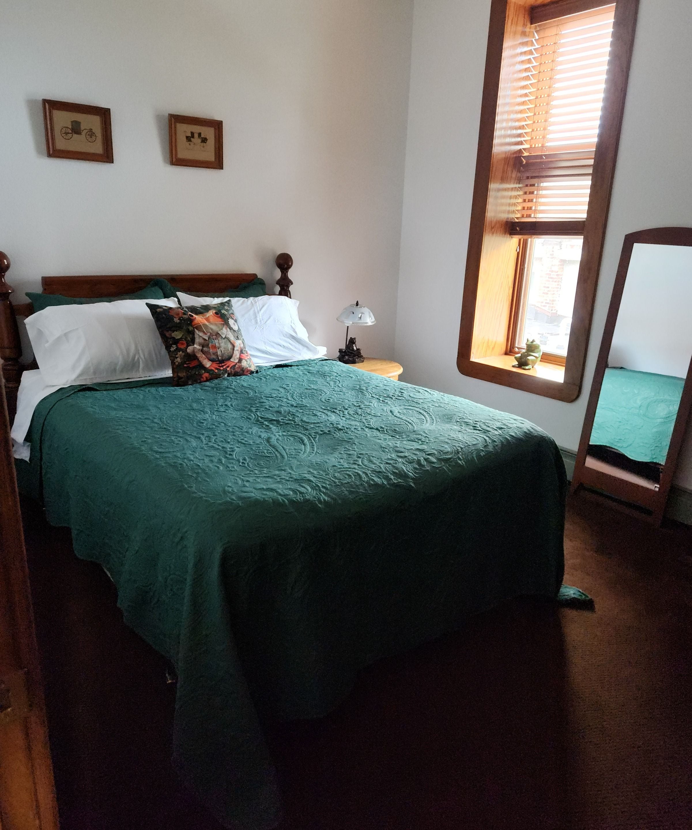 A bed with a green bedspread, a wooden headboard, a nightstand with a lamp, and a window with blinds.