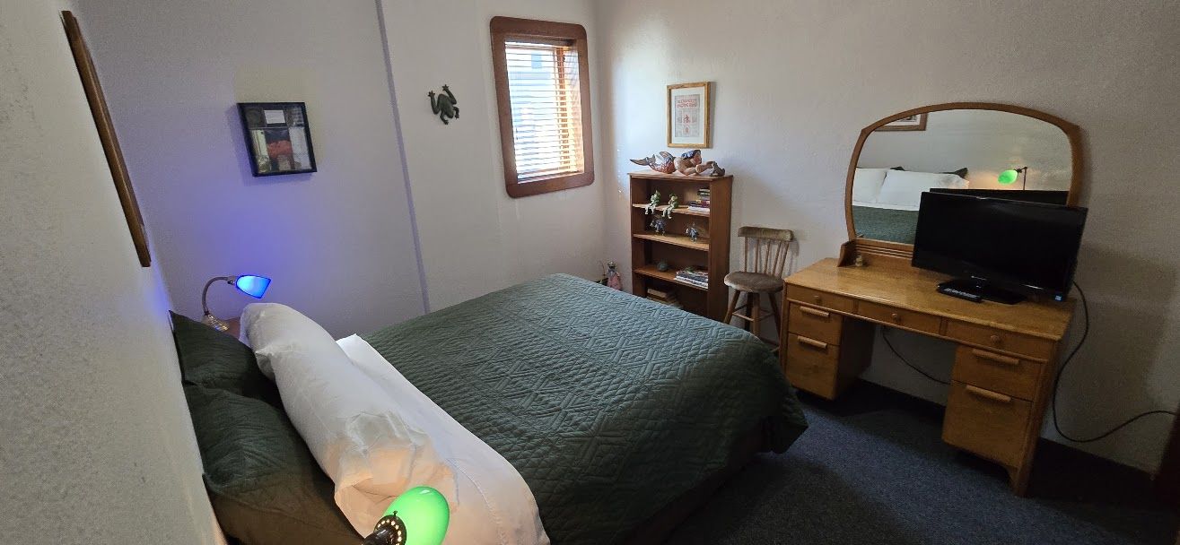 A bedroom with a bed, a desk with a computer, a chair, a bookshelf, and a window with blinds.