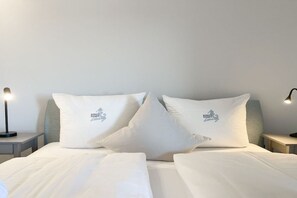 Een hotelkamer met een net opgemaakt bed, twee kussens met een logo, een nachtkastje met een lamp en een nachttafeltje.