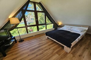 A bedroom with a sloped ceiling, a bed, a TV, and a window with a view of greenery. - Holiday home with 3 bedrooms (Sierksdorf)