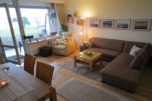 Una sala de estar con sofá, sillón, mesa de centro y mesa de comedor. Hay una ventana con cortinas, una lámpara y cuadros en la pared.