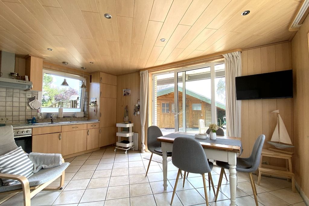 A modern kitchen with wooden cabinets, a dining area with a table and chairs, and a TV mounted on the wall.