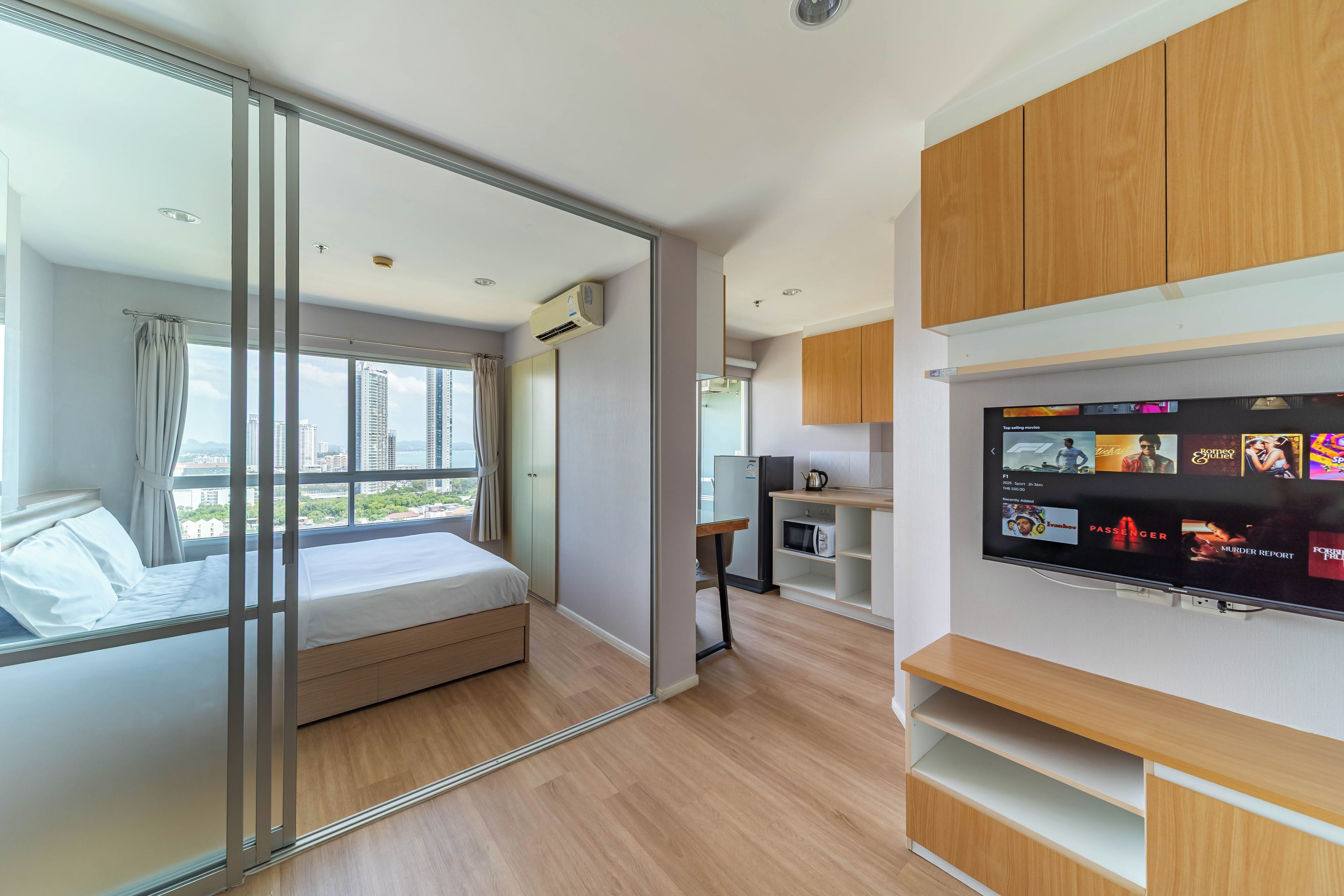 A modern hotel room with a bed, a TV, a kitchenette, and a sliding glass door to a balcony.