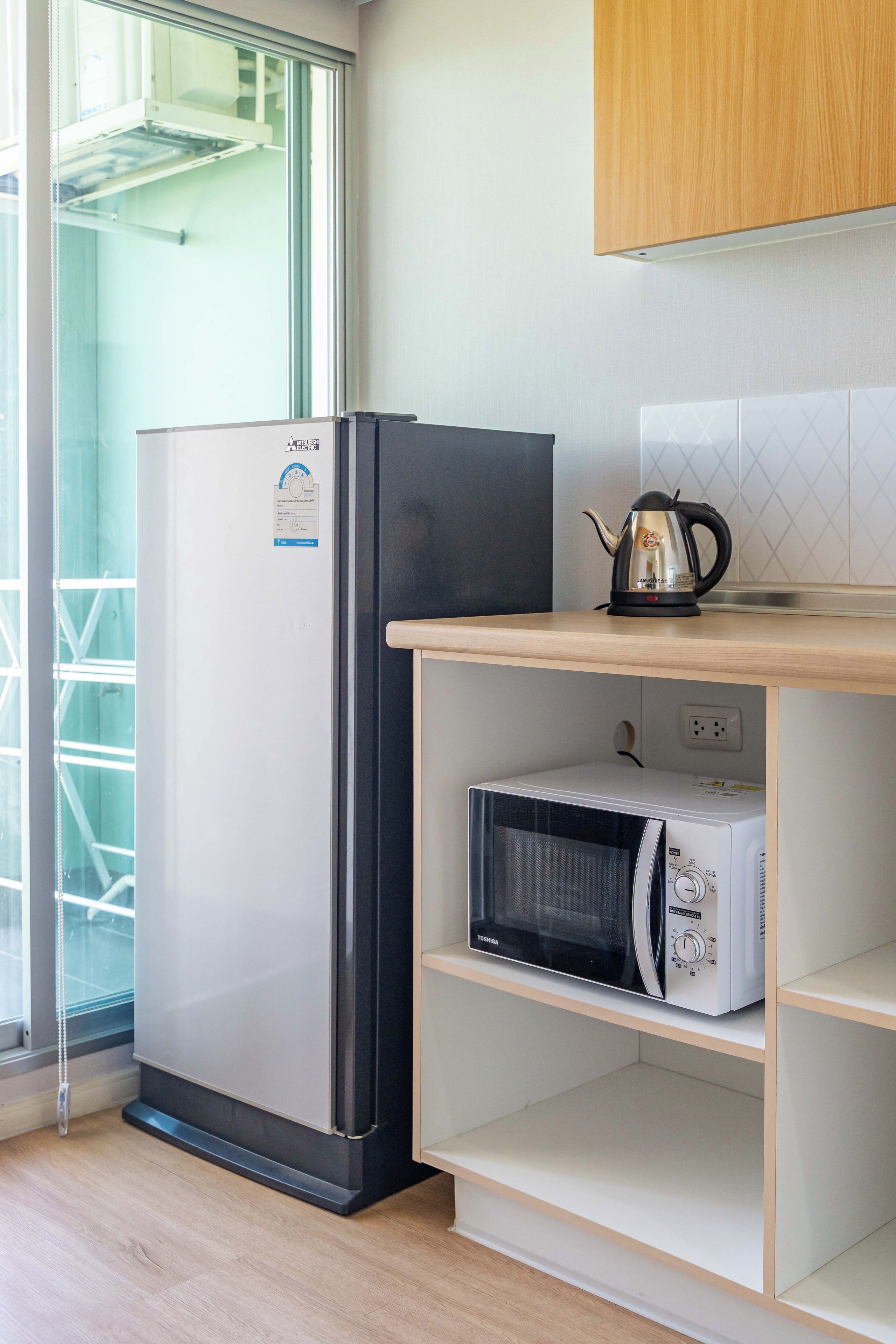A kitchen with a refrigerator, microwave, kettle, and wooden cabinets.