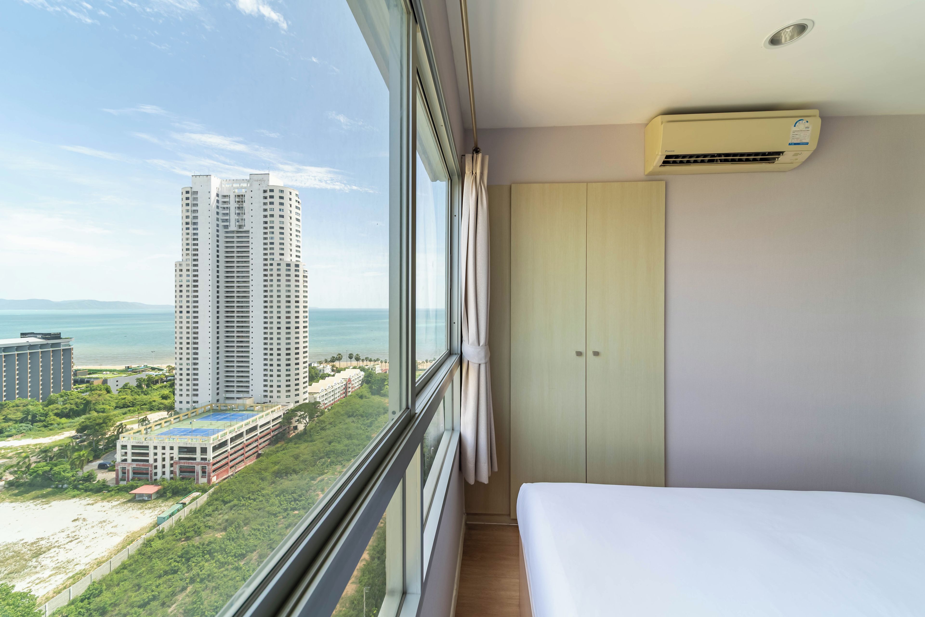A hotel room with a large window, a bed, a wardrobe, and an air conditioning unit.
