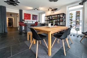 A modern dining area with a wooden table and black chairs, a bookshelf, and a view of the outside through a large window. - resin passion || Braunlage (Braunlage)