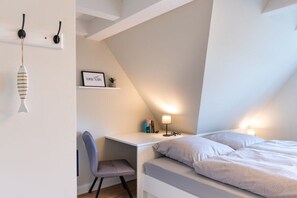Un dormitorio con cama, escritorio, silla, estante con libros y un cuadro, y una lámpara de pared.