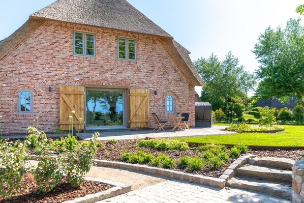 A brick house with a thatched roof, a wooden door, and a garden with plants and a paved area.