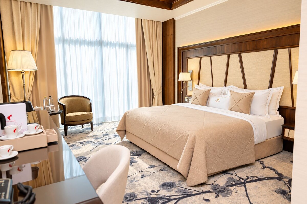 Quarto de hotel com cama grande, escrivaninha com telefone, cadeira e vista do exterior através de cortinas translúcidas.