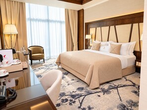Quarto de hotel com cama grande, escrivaninha com telefone, cadeira e vista do exterior através de cortinas translúcidas.