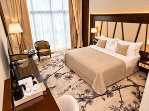 Quarto de hotel com uma cama grande, escrivaninha com cafeteira, cadeira e janela com cortinas.