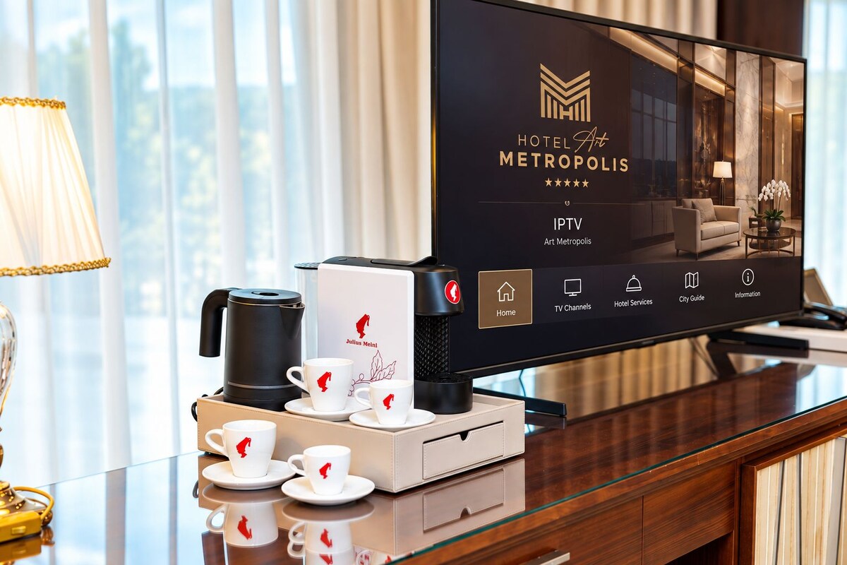 Quarto de hotel com tela digital exibindo o nome e os serviços do hotel, uma cafeteira e um conjunto de xícaras brancas com um logotipo vermelho.