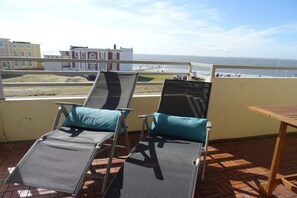 Ein Balkon mit zwei Liegestühlen und einem Tisch, mit Blick auf einen Strand und Gebäude.