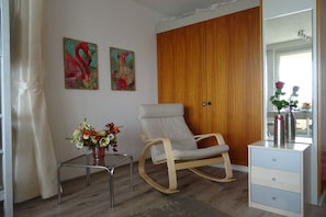Ein Zimmer mit einem Holzschrank, einem weißen Stuhl, einem kleinen Tisch mit Blumenvase und zwei an der Wand befestigten Bildern.