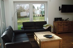 Un salon avec un canapé noir, une table basse en bois, un téléviseur fixé au mur et un meuble contenant une radio.
