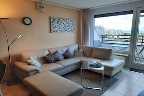 A modern living room with a beige sectional sofa, a glass coffee table, a floor lamp, and a city view through large windows. - Holiday apartment in Damp with swimming pool (Damp)