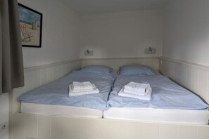 Un lit simple avec deux oreillers et des serviettes pliées, des murs blancs et un tableau encadré accroché au mur.