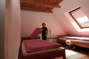 Ein Zimmer mit zwei hölzernen Betten, eines mit einer rot karierten Bettdecke und das andere mit einer weißen. Über den Betten befindet sich ein Dachfenster, und in der Ecke ist ein kleiner Tisch mit einer Lampe zu sehen.