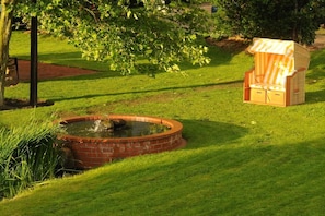 Ein Garten mit einem runden Backsteinbrunnen, einer hölzernen Bank und einem gestreiften Gartenstuhl.