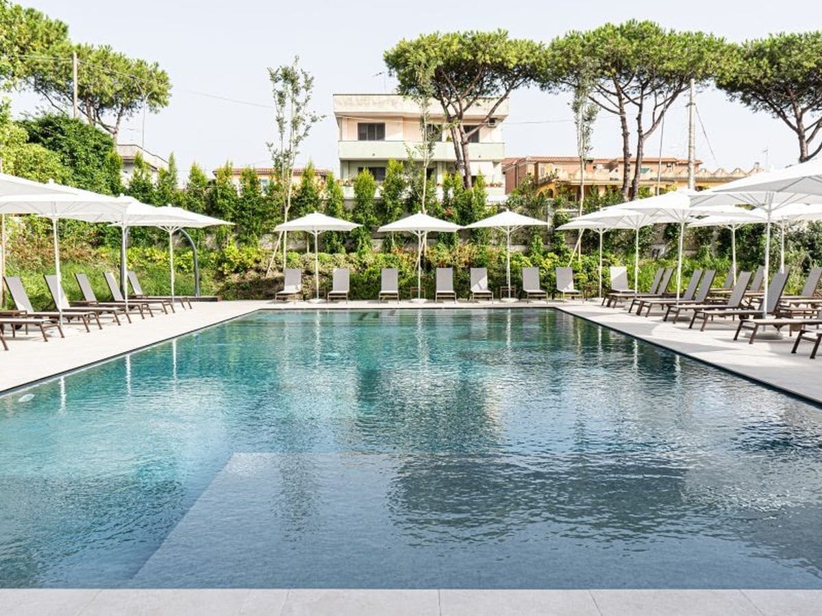 Una piscina rectangular con sillas de descanso y sombrillas, rodeada de follaje y edificios al fondo.