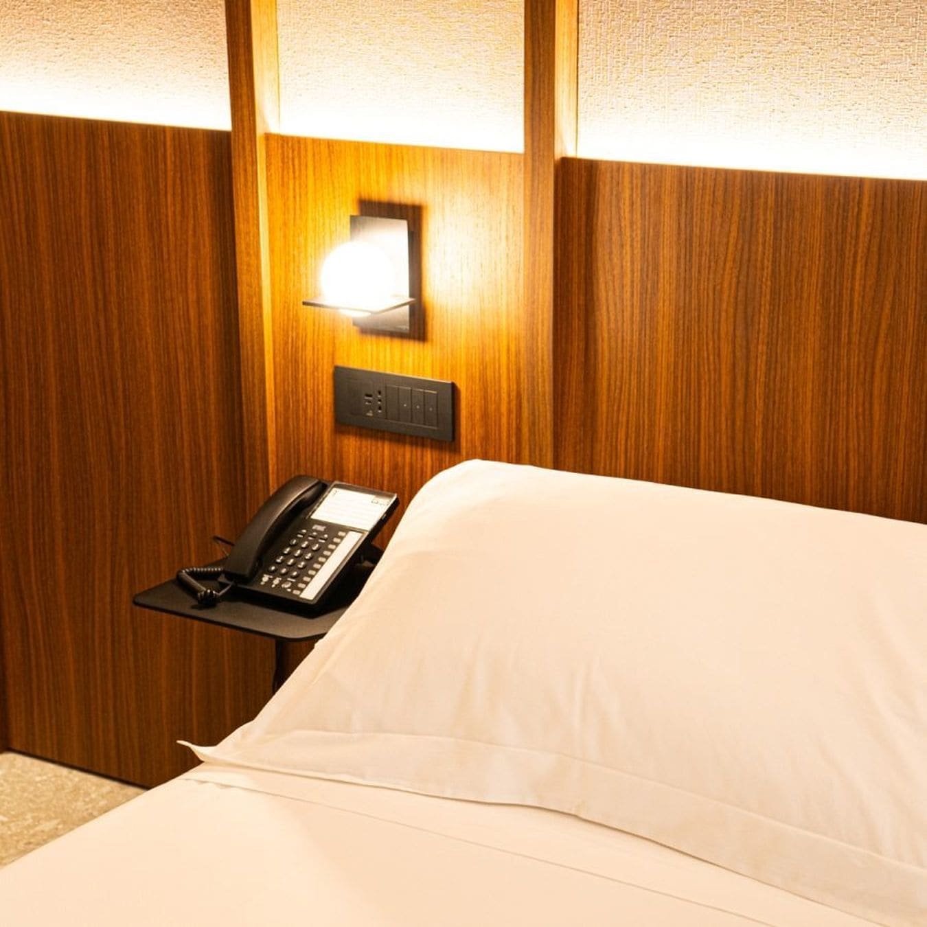 Una camera d'albergo con un letto, un telefono sul comodino e un punto luce a parete.