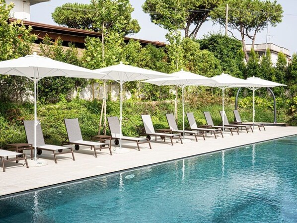 A poolside area with lounge chairs and umbrellas, surrounded by greenery and a building in the background.