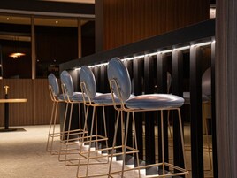 Una hilera de taburetes de bar con estructura metálica y asientos acolchados, ubicados frente a una barra de madera oscura.