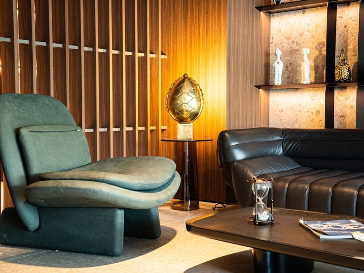 Un espace de détente moderne avec un fauteuil vert, un canapé en cuir noir et une petite table sur laquelle se trouvent une lampe et un sablier.