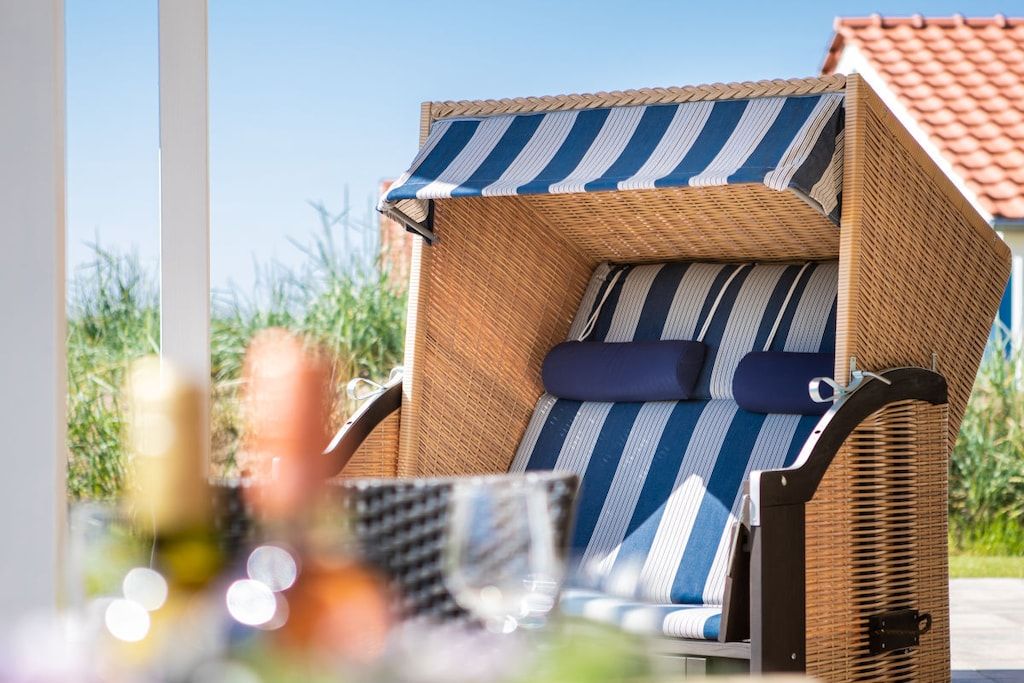 A wicker lounge chair with striped cushions, positioned outdoors with a view of greenery and a building in the background.