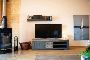 A living room with a flat-screen TV, wooden flooring, a fireplace, and a wall-mounted shelf with books and decorative items. - "P-Liner" house type E plus (Neustadt\/Holstein - OT Pelzerhaken)